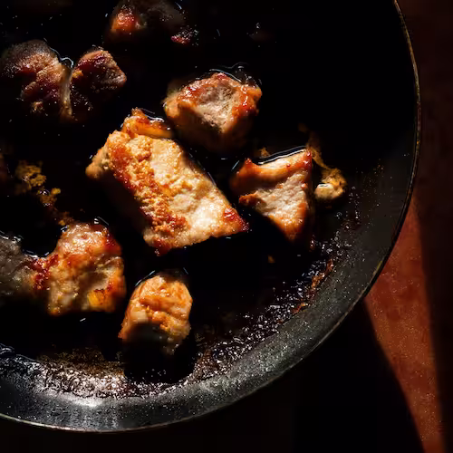 Pork searing in a hot pan with oil crackling around meat forming golden-brown caramelization.